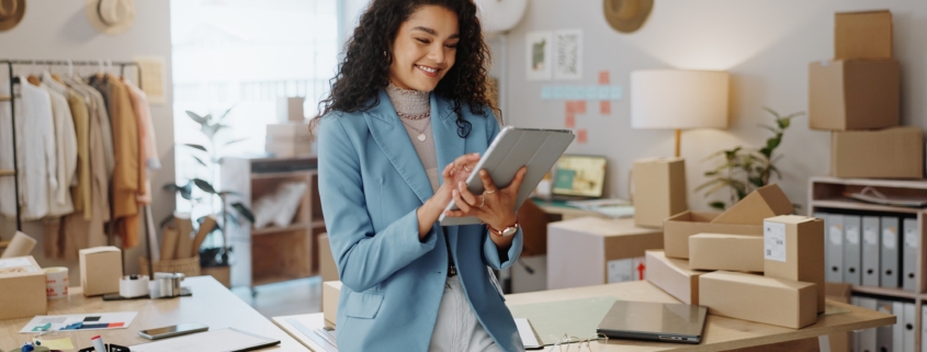 A woman leans on a table in a small retail space. She's looking down at a tablet she's holding.