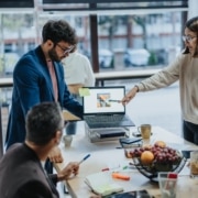 A,Diverse,Group,Of,Professionals,Collaborate,In,A,Modern,Office, A diverse group of professionals collaborate in a modern office. They stand around a table looking at a presentation on a laptop.