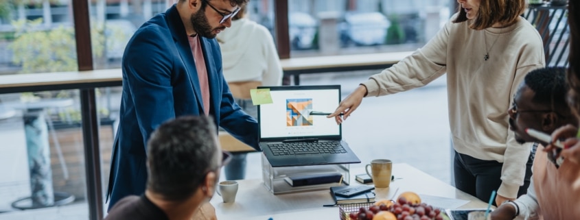 A diverse group of professionals collaborate in a modern office. They stand around a table looking at a presentation on a laptop.
