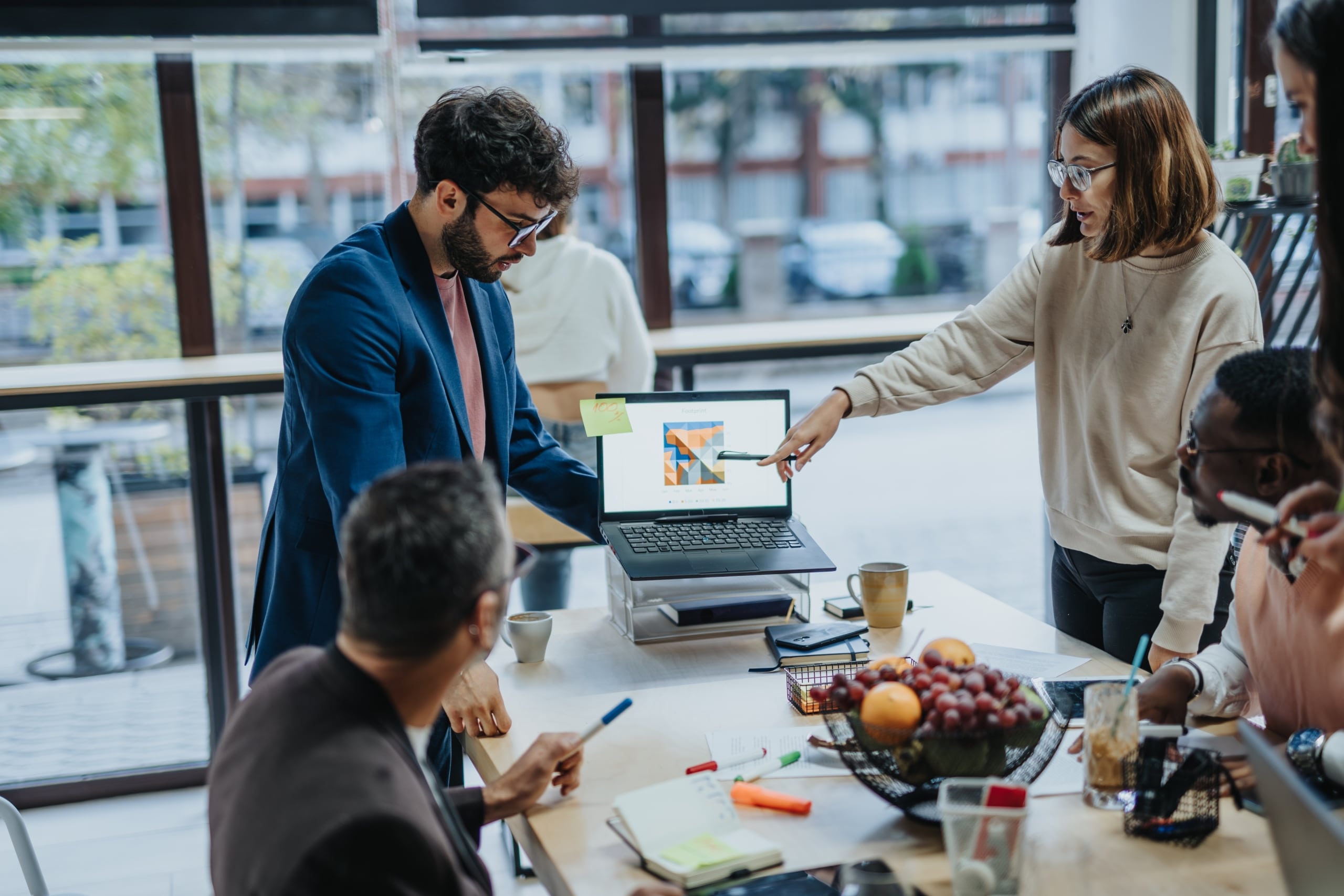 A diverse group of professionals collaborate in a modern office. They stand around a table looking at a presentation on a laptop.