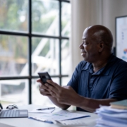 A Black man sits at a desk with a laptop and stacks of paper around him. He holds a phone while looking out of the window. The image shows capacity planning before growing your business.