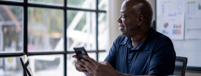 A Black man sits at a desk with a laptop and stacks of paper around him. He holds a phone while looking out of the window. The image shows capacity planning before growing your business.
