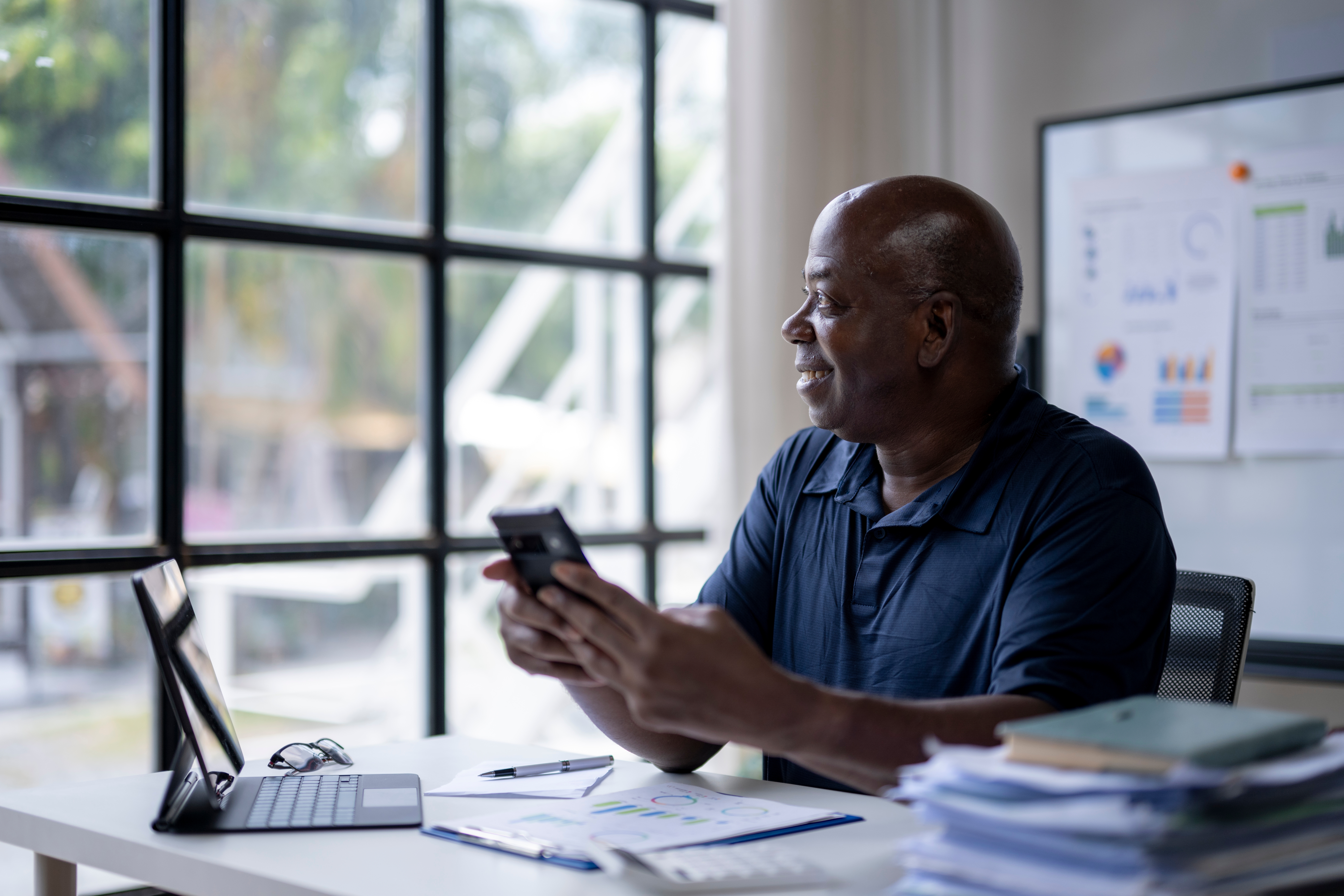 A Black man sits at a desk with a laptop and stacks of paper around him. He holds a phone while looking out of the window. The image shows capacity planning before growing your business.