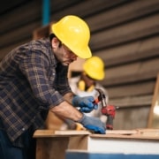 Focused,Craftsman,In,An,Industrial,Workshop,,Demonstrating,Woodworking,Techniques,And A man wearing a yellow hardhat and work gloves hammers a nail into a wooden board.