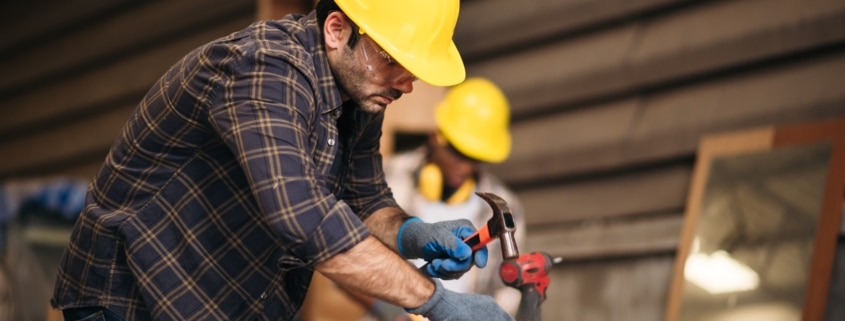 A man wearing a yellow hardhat and work gloves hammers a nail into a wooden board.