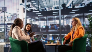 Three women are in discussion around a table in an office building.