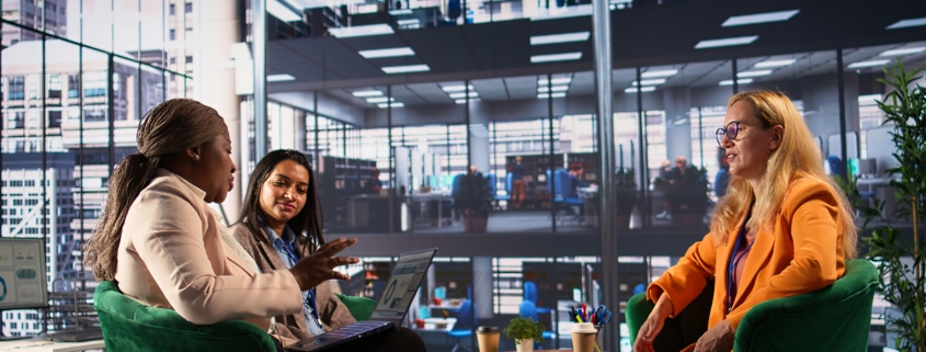 Three women are in discussion around a table in an office building.