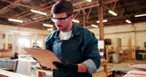 A man wearing work overalls, safety glasses, and work gloves checks items off a checklist on a clipboard.