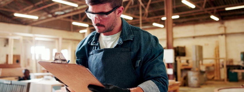 A man wearing work overalls, safety glasses, and work gloves checks items off a checklist on a clipboard.