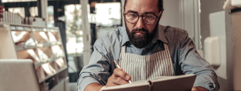 A small business owner wonders why lenders cap financing amounts. A man sits at a desk with a computer in front of him, he's writing in a notebook looking concerned.