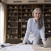 A female small business owner contemplates how lenders assess risk in small business financing. She stands at a workbench in a studio with an open laptop and papers in front of her.