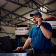 Mature,Mechanic,Communicating,On,Cell,Phone,While,Working,On,Laptop Auto mechanic talking on the phone in his repair garage while researching how to prepare for a business loan for his small business.