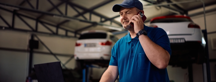 Auto mechanic talking on the phone in his repair garage while researching how to prepare for a business loan for his small business.